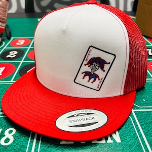 Op de afbeelding: Een witte en rode baseballpet met een mesh achterkant. De voorkant van de pet heeft een geborduurd beeld van een joker speelkaart. De pet heeft een zwart label met de tekst "THE AUTHENTIC YUPOONG SNAPBACK YUPOONG SINCE 1974".