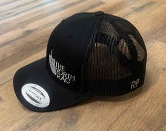 Może przedstawiać: Czarna i szara truckerka marki The North Face z siateczką z tyłu i białym logo "The North Face". Czapka ma czarne zapięcie na zatrzask i białą naklejkę "The Authentic Snapback" na spodzie daszka.