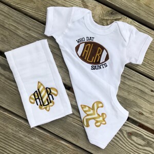 Puede incluir: Body de beb&eacute; blanco y pa&ntilde;o de eructo a juego con un dise&ntilde;o de f&uacute;tbol americano. El body tiene el texto "WHO DAT" y "SAINTS". El bal&oacute;n de f&uacute;tbol es marr&oacute;n con letras doradas. El pa&ntilde;o de eructo y el body tambi&eacute;n tienen un dise&ntilde;o de flor de lis dorada.
