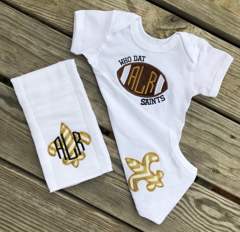 Puede incluir: Body de beb&eacute; blanco y pa&ntilde;o de eructo a juego con un dise&ntilde;o de f&uacute;tbol americano. El body tiene el texto "WHO DAT" y "SAINTS". El bal&oacute;n de f&uacute;tbol es marr&oacute;n con letras doradas. El pa&ntilde;o de eructo y el body tambi&eacute;n tienen un dise&ntilde;o de flor de lis dorada.