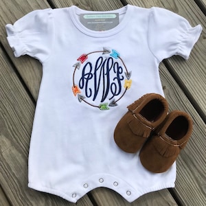 May include: White baby romper with a navy blue monogram encircled by colourful arrows. The romper has short sleeves and popper fastenings. Brown leather baby moccasins with fringe are next to the romper. The text "Southern Grace Embroidery" is on the top.