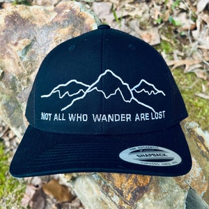 May include: Black trucker hat with a white embroidered mountain range design and the text "Not all who wander are lost". The hat has a black mesh back and a black snap closure.