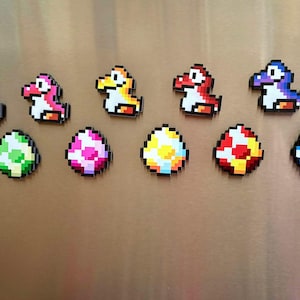 May include: A set of eight magnets featuring pixelated characters and eggs from the Super Mario Bros. video game. The characters are Yoshi in green, pink, yellow, and purple. The eggs are green, pink, yellow, red, and blue.