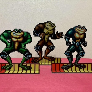 Puede incluir: Tres figuras pixeladas de ranas verdes, marrones y azules de pie en una plataforma amarilla y roja. Las figuras son de un videojuego.