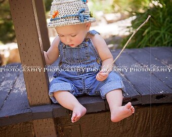 May include: A young child wearing a crocheted hat with colorful flowers and blue overalls sits on a wooden bridge holding a stick fishing rod. The fishing line is in the water with a hook.