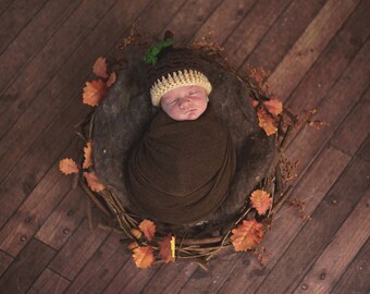 Puede incluir: Un bebé recién nacido envuelto en una manta marrón, con un gorro de bellota de ganchillo con un tallo verde y un borde color crema. El bebé descansa en un nido de ramitas y hojas de otoño, sobre un fondo de madera.