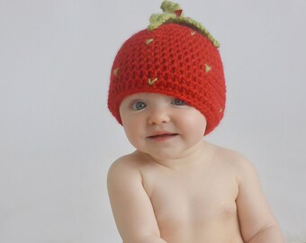 May include: A baby wearing a red crocheted strawberry hat with green leaves. The hat has yellow stitching details.
