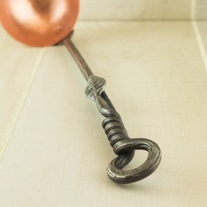 Op de afbeelding: Een koperen lepel met een lange, gedraaide, ijzeren steel. De steel is versierd met een bladontwerp.