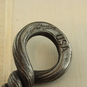 Op de afbeelding: Close-up van een gedraaide metalen haak met een ring aan het einde. De ring is gestempeld met "USA" en een logo. De haak is gemaakt van donker metaal.