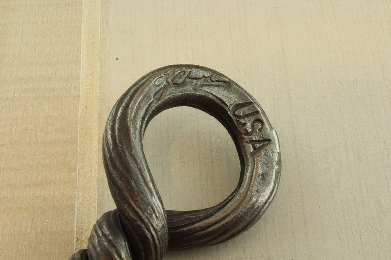 Op de afbeelding: Close-up van een gedraaide metalen haak met een ring aan het einde. De ring is gestempeld met "USA" en een logo. De haak is gemaakt van donker metaal.