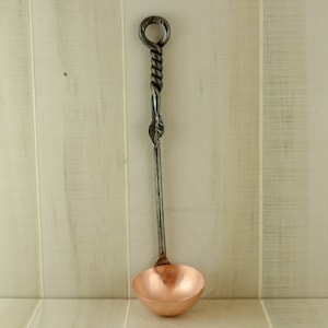 Op de afbeelding: Een koperen lepel met een lange, gedraaide, zwarte metalen steel. De steel heeft een lus aan de bovenkant.