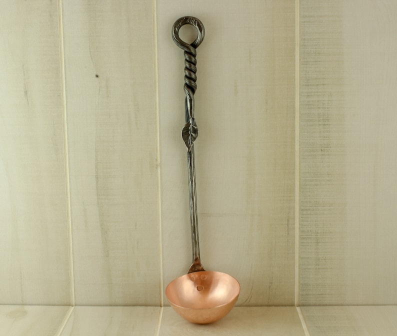 Op de afbeelding: Een koperen lepel met een lange, gedraaide, zwarte metalen steel. De steel heeft een lus aan de bovenkant.