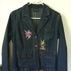 May include: A dark blue denim jacket with two embroidered fairy patches. The jacket has a button closure and two front pockets.