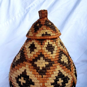 May include: A large, woven basket with a lid. The basket is brown, black, and beige and has a pear-shaped body. The lid is a dome shape with a pointed top.