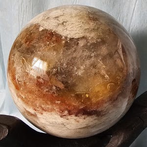 May include: A large, round, clear quartz crystal sphere with brown, white, and yellow inclusions. The sphere is polished and has a smooth surface.