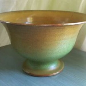 May include: A green and brown ceramic bowl with a pedestal base. The bowl has a glazed finish and a slightly flared rim.
