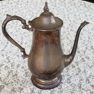 May include: An antique silver-colored coffee pot with a curved handle and a long, slender spout. The pot has a lid with a decorative finial and sits on a round base. The metal shows signs of age and use.