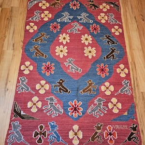 May include: A rectangular, hand-woven rug with a red background and a central blue diamond design. The rug features repeating patterns of stylized animals, flowers, and birds in various colors, including brown, gray, and yellow. The rug has a fringed edge.
