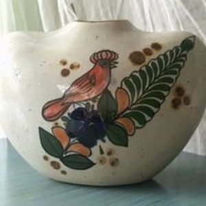 May include: Cream-colored ceramic vase with a wide, curved shape. The vase features a hand-painted design of a red bird, green leaves, and blue flowers. Brown spots and orange accents complete the nature-inspired artwork. The vase is approximately 8 inches wide.