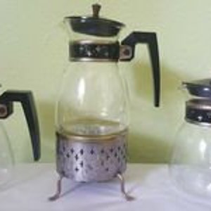 May include: Set of three glass coffee pots with black lids and handles. The center pot has a metal stand with three legs.