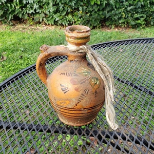 May include: A brown ceramic jug with a handle and a white rope tied around the neck. The jug has a painted design of flowers and leaves in shades of orange, yellow, and green. The jug is sitting on a black metal table with a diamond-shaped pattern.