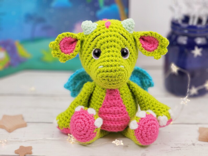 Puede incluir: Un drag&oacute;n de crochet verde con detalles rosas y azules. El drag&oacute;n tiene ojos grandes y una barriga rosa. Est&aacute; sentado sobre una superficie blanca.