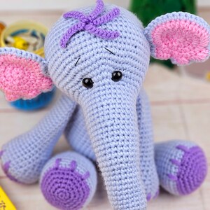 May include: A crocheted elephant toy with a light blue body, pink ears, and purple accents. The elephant has black eyes and a long, gray trunk.