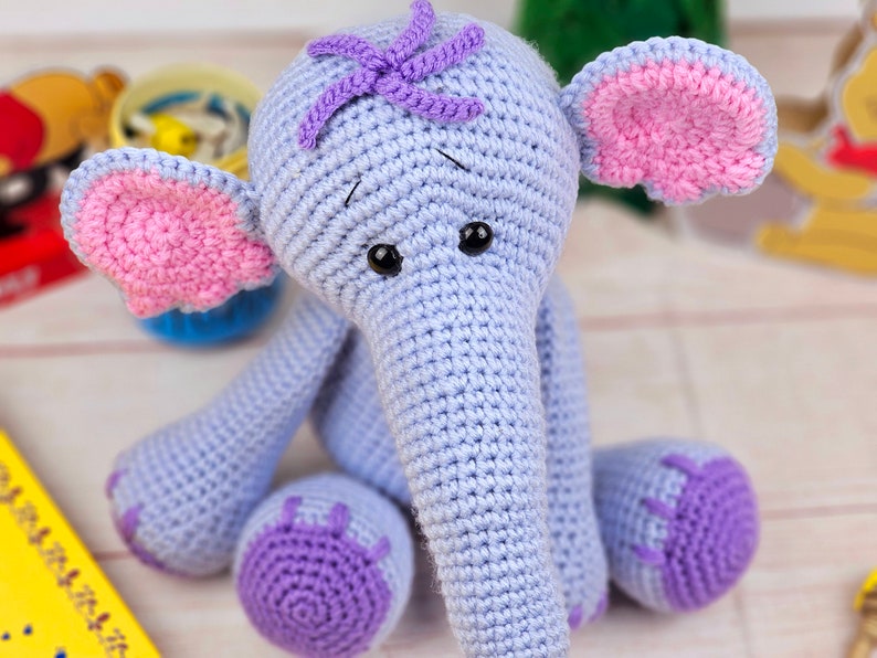 May include: A crocheted elephant toy with a light blue body, pink ears, and purple accents. The elephant has black eyes and a long, gray trunk.