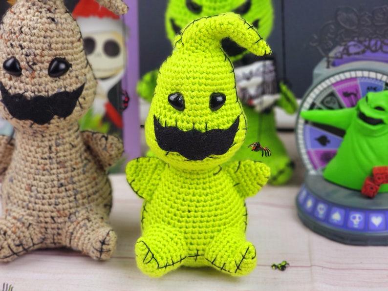 Puede incluir: Un juguete de peluche verde tejido a crochet del personaje Oogie Boogie de la pel&iacute;cula Pesadilla antes de Navidad. El juguete tiene una boca cosida negra, ojos negros y un bigote cosido negro. El juguete est&aacute; sentado sobre una superficie blanca.