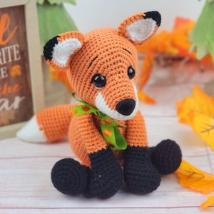 May include: A crocheted orange fox with black feet and a green and orange ribbon around its neck. The fox is sitting on a white surface with fall leaves in the background.