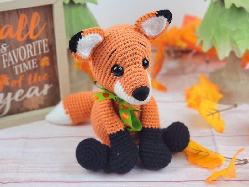 May include: A crocheted orange fox with black feet and a green and orange ribbon around its neck. The fox is sitting on a white surface with fall leaves in the background.