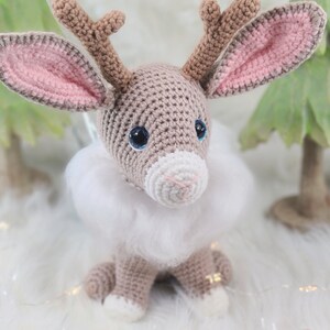 May include: A crocheted reindeer figurine with large blue eyes, brown antlers, and pink-lined ears. It has a white fluffy collar and is sitting on a white surface. The reindeer is a handmade item, perfect for holiday decor.