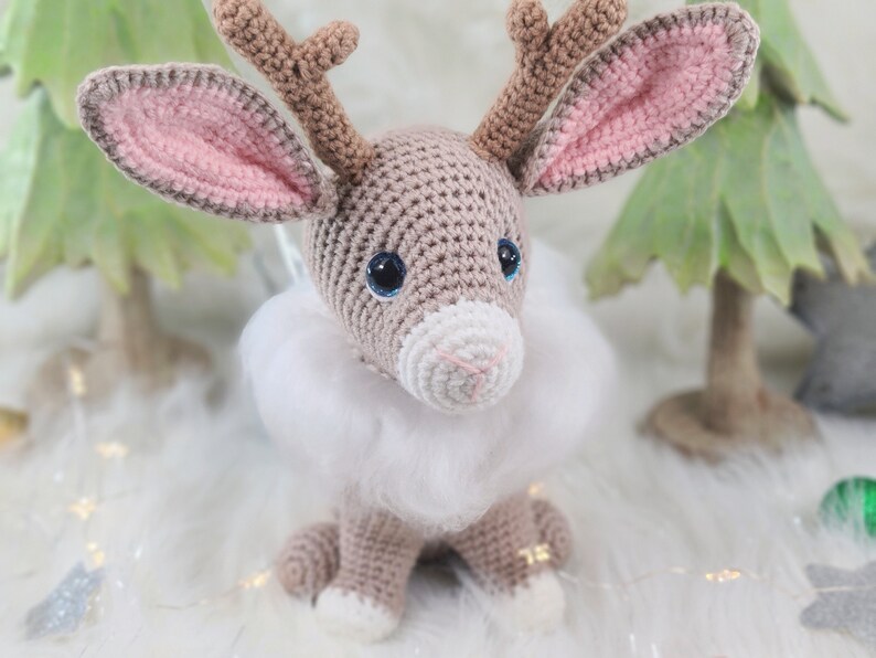 May include: A crocheted reindeer figurine with large blue eyes, brown antlers, and pink-lined ears. It has a white fluffy collar and is sitting on a white surface. The reindeer is a handmade item, perfect for holiday decor.