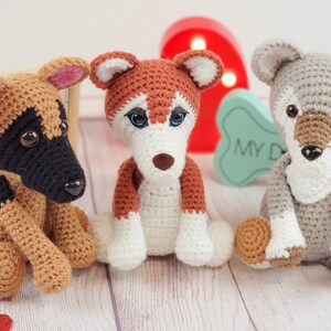 May include: Three crocheted stuffed dog toys. The dog on the left is brown and black, the middle dog is brown and white, and the dog on the right is gray. The dogs are sitting on a light wood surface.