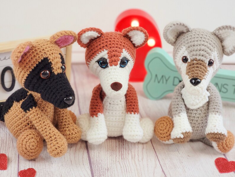 May include: Three crocheted stuffed dog toys. The dog on the left is brown and black, the middle dog is brown and white, and the dog on the right is gray. The dogs are sitting on a light wood surface.
