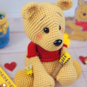 K&ouml;nnte beinhalten: Ein geh&auml;keltes Winnie Puuh-B&auml;ren-Spielzeug, das ein rotes Hemd tr&auml;gt und einen Honigl&ouml;ffel h&auml;lt. Der B&auml;r ist gelb und braun und sitzt auf einer wei&szlig;en Oberfl&auml;che.