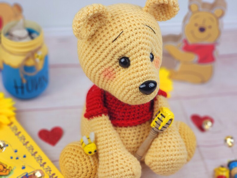 K&ouml;nnte beinhalten: Ein geh&auml;keltes Winnie Puuh-B&auml;ren-Spielzeug, das ein rotes Hemd tr&auml;gt und einen Honigl&ouml;ffel h&auml;lt. Der B&auml;r ist gelb und braun und sitzt auf einer wei&szlig;en Oberfl&auml;che.