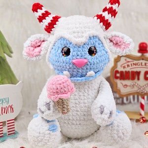 Pu&ograve; includere: Un giocattolo di peluche bianco all'uncinetto con pelliccia blu sul viso e un cono gelato rosa in mano. Il giocattolo ha corna a strisce rosse e bianche ed &egrave; seduto davanti a un cartello che dice "Kringle & Co. Candy Canes".