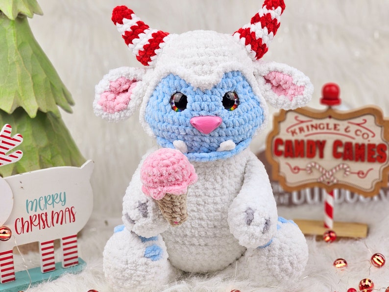 Pu&ograve; includere: Un giocattolo di peluche bianco all'uncinetto con pelliccia blu sul viso e un cono gelato rosa in mano. Il giocattolo ha corna a strisce rosse e bianche ed &egrave; seduto davanti a un cartello che dice "Kringle & Co. Candy Canes".