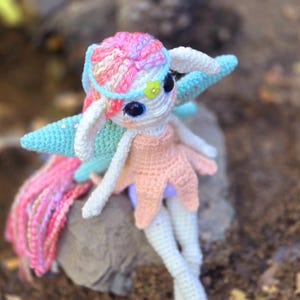 May include: A crocheted fairy doll with pink, blue, and white yarn. The doll has a long, flowing pink and white tail and is wearing a pink dress with a blue and white flower on its head.