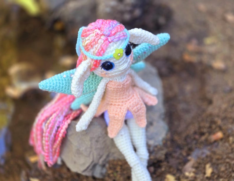 May include: A crocheted fairy doll with pink, blue, and white yarn. The doll has a long, flowing pink and white tail and is wearing a pink dress with a blue and white flower on its head.