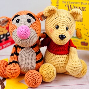May include: Two crocheted stuffed animals, Tigger and Winnie the Pooh, sitting together. Tigger is orange and black with a pink nose. Winnie the Pooh is yellow with a red shirt.