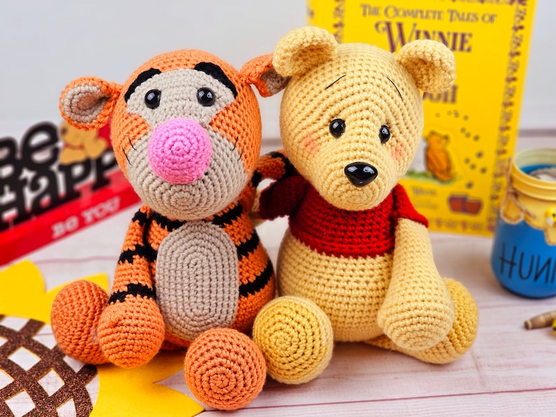 May include: Two crocheted stuffed animals, Tigger and Winnie the Pooh, sitting together. Tigger is orange and black with a pink nose. Winnie the Pooh is yellow with a red shirt.