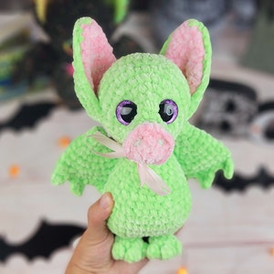 May include: A green crocheted bat plush toy with pink ears and a pink nose. The toy has large purple eyes and a white ribbon around its neck.