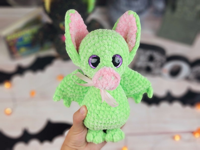May include: A green crocheted bat plush toy with pink ears and a pink nose. The toy has large purple eyes and a white ribbon around its neck.