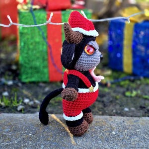 Puede incluir: Un juguete de peluche de crochet con forma de criatura negra y gris que lleva un gorro de Pap&aacute; Noel rojo y blanco y un atuendo rojo y blanco. El juguete est&aacute; de pie sobre una superficie gris.