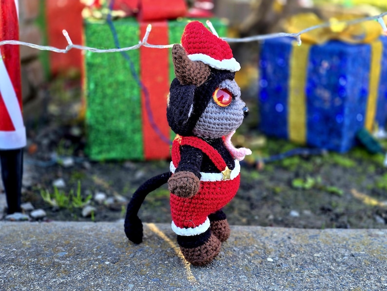 Puede incluir: Un juguete de peluche de crochet con forma de criatura negra y gris que lleva un gorro de Pap&aacute; Noel rojo y blanco y un atuendo rojo y blanco. El juguete est&aacute; de pie sobre una superficie gris.