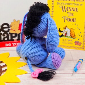 May include: A blue crocheted Eeyore plush toy with a black mane and a pink tail. The toy is sitting on a wooden surface with a sunflower and a book titled "The Complete Tales of Winnie the Pooh" in the background.