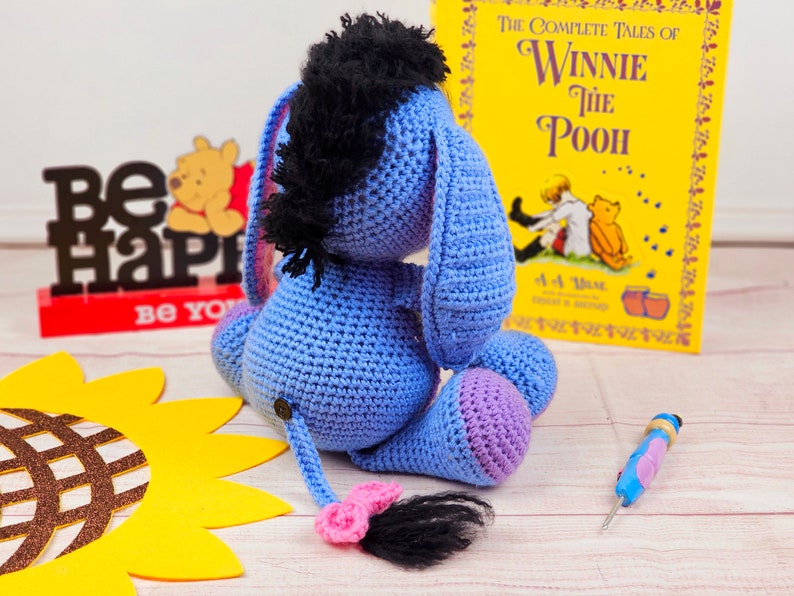 May include: A blue crocheted Eeyore plush toy with a black mane and a pink tail. The toy is sitting on a wooden surface with a sunflower and a book titled "The Complete Tales of Winnie the Pooh" in the background.