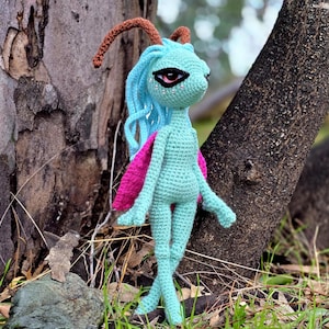 May include: A crocheted blue and pink fairy doll with long blue hair and a brown antenna. The doll has a single black eye and is standing in front of a tree trunk.